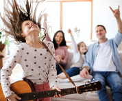 Image of a 12-year-old girl energetically strumming her guitar takes center stage, while in the background, her family shares hearty laughter while gathered on a sofa.